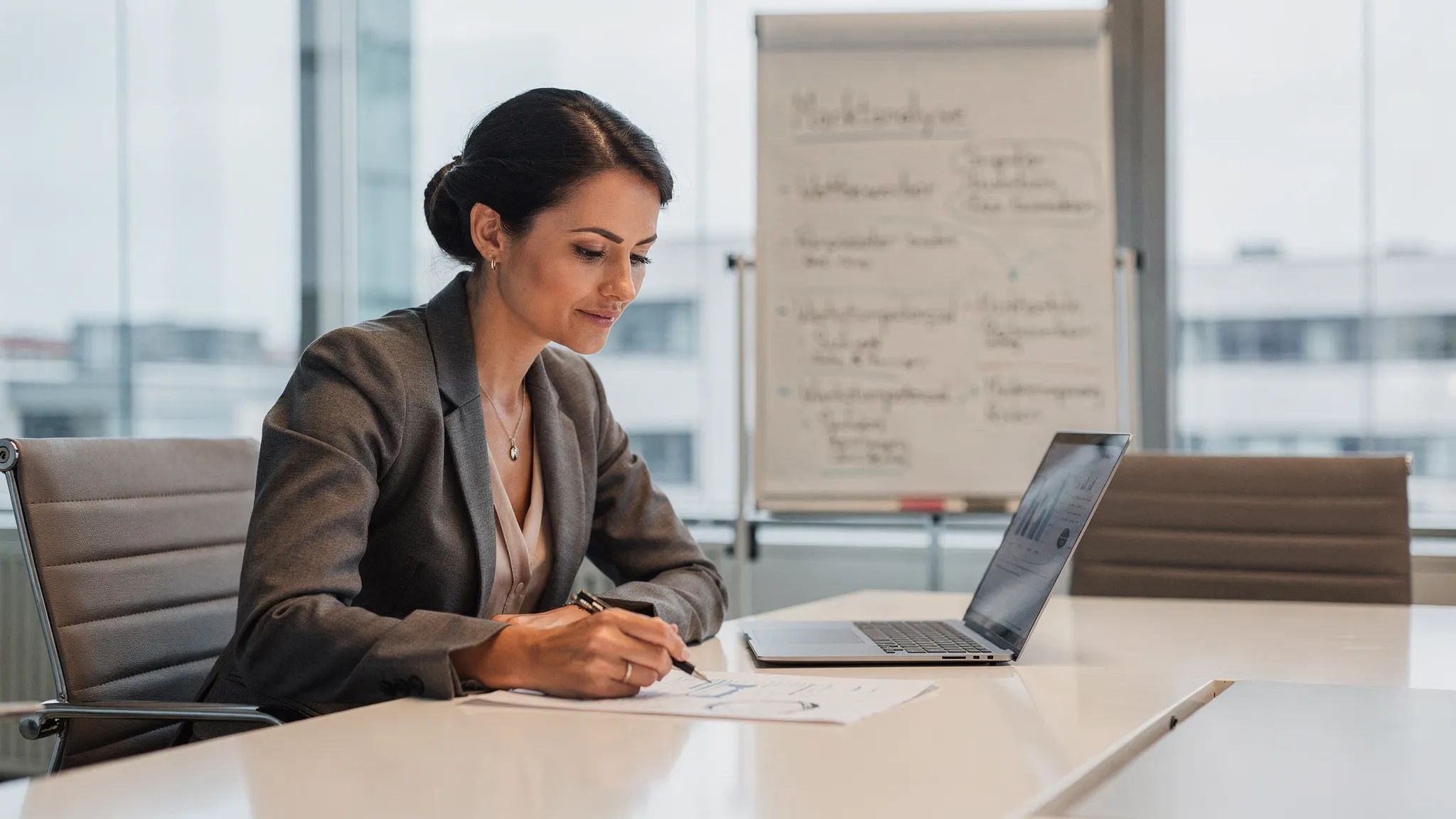Eine Analystin in einem Besprechungsraum prüft einen Due-Diligence-Bericht auf Papier, daneben ein Laptop (Bildschirm nach rechts ausgerichtet, ohne sichtbare vertrauliche Inhalte), im Hintergrund ein Whiteboard mit Markt- und Wettbewerbsnotizen.